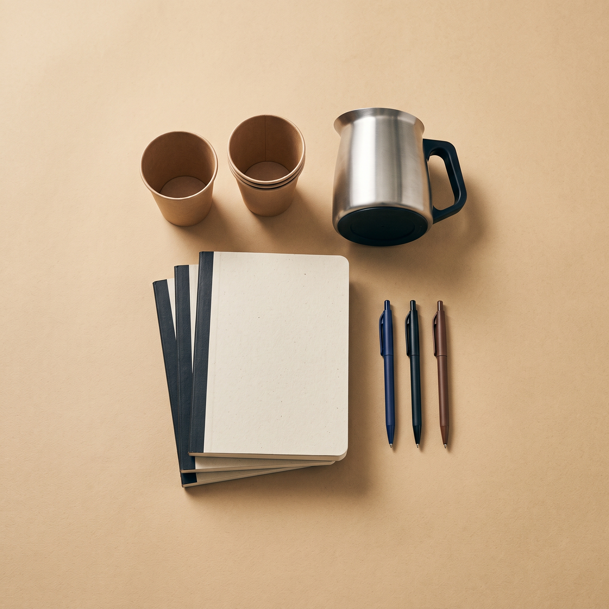 Notebooks, pens, a coffee carafe and cups on a sand-colored surface